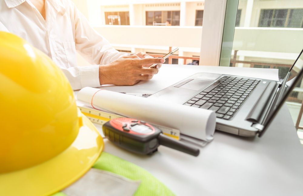 Construction professional reviewing blueprints and smartphone next to laptop, hard hat and walkie‑talkie on desk