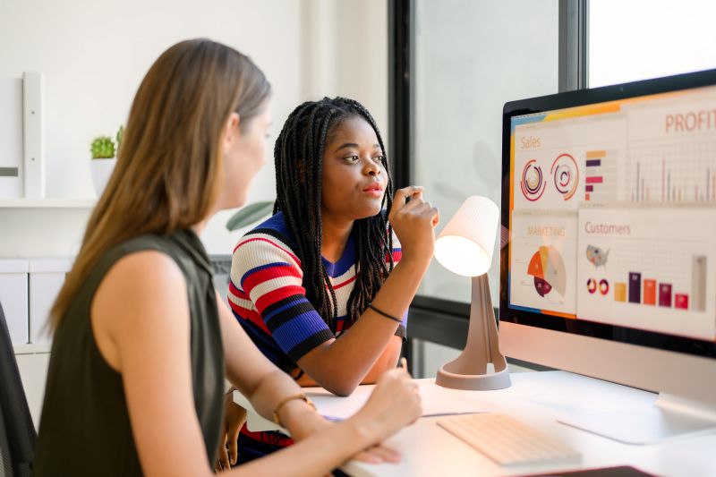 Deux femmes en réunion face à un écran affichant des graphiques de ventes, marketing et clients, diagrammes circulaires et barres colorées.